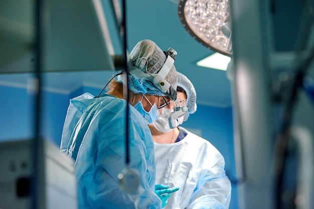 Chirurgien effectuant une chirurgie esthétique dans la salle d'opération de l'hôpital Chirurgien en masque portant des loupes pendant la procédure médicale Amélioration de l'élargissement de l'augmentation mammaire Filtre bleu fond bleu