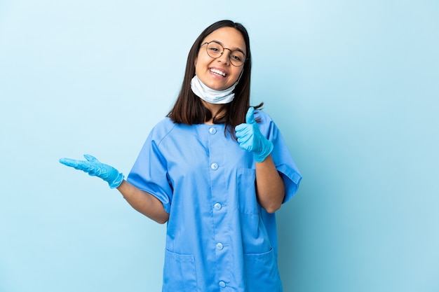 Chirurgien femme sur mur bleu isolé tenant copyspace imaginaire sur la paume pour insérer une annonce et avec les pouces vers le haut