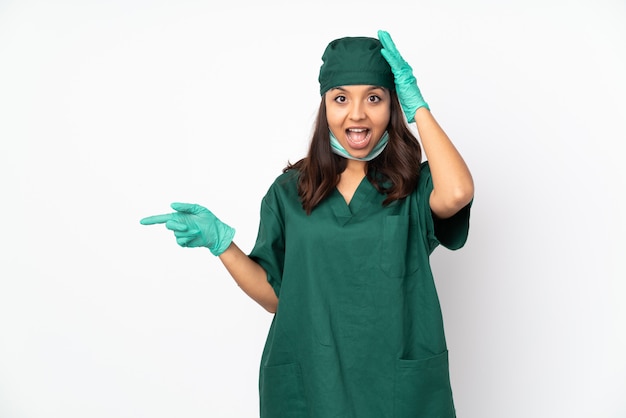 Chirurgien femme en uniforme vert sur mur blanc surpris et pointant le doigt sur le côté