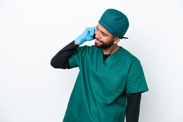 Chirurgien homme brésilien en uniforme vert isolé sur fond blanc en gardant une conversation avec le téléphone mobile avec quelqu'un