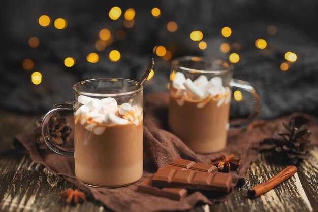 Chocolat chaud d'hiver riche avec des bâtons de cannelle et des barres de chocolat aux noix dans une tasse transparente sur une planche en bois mise au point sélective avec une guirlande festive