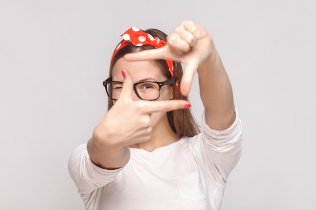 Choisir la composition ou se concentrer sur le portrait d'une belle jeune femme émotive en t-shirt blanc avec des taches de rousseur, des lunettes, des lèvres rouges et un bandeau. tourné en studio intérieur, isolé sur fond gris clair.