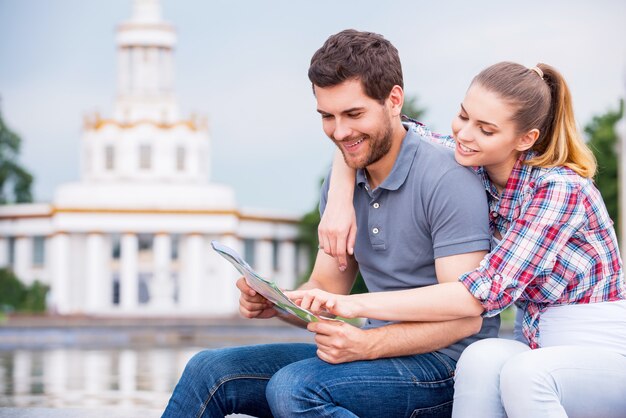 Choisir l'endroit où aller. Heureux jeune couple de touristes assis près d'un beau bâtiment et examinant la carte ensemble