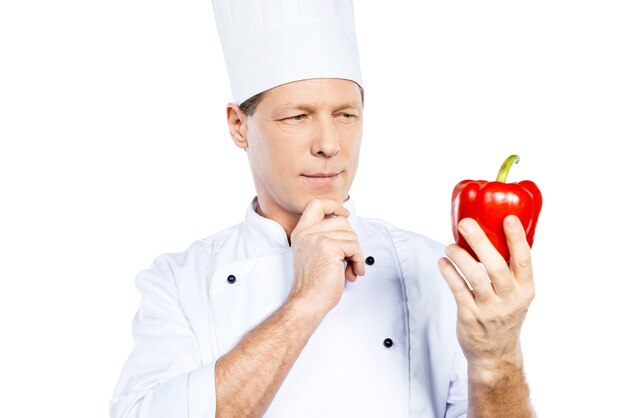 Choisir les meilleurs légumes pour son repas. Cheerful mature chef en uniforme blanc tenant du poivron rouge et le regardant en se tenant debout sur fond blanc