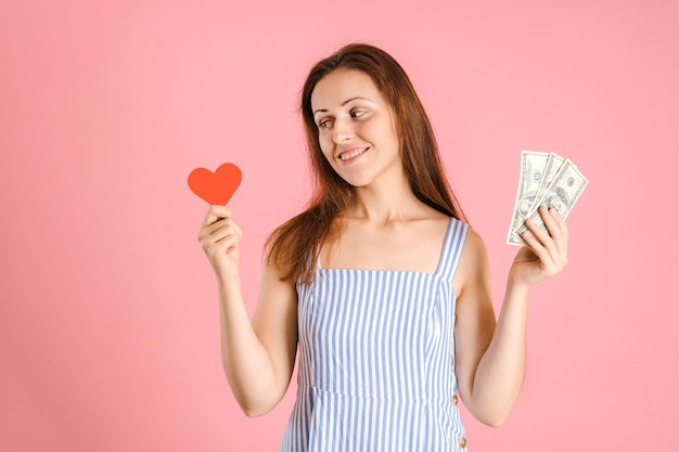 Le choix entre l'amour et l'argent. Une femme tient de l'argent et un symbole de coeur rouge dans ses mains. Studio tourné sur fond rose