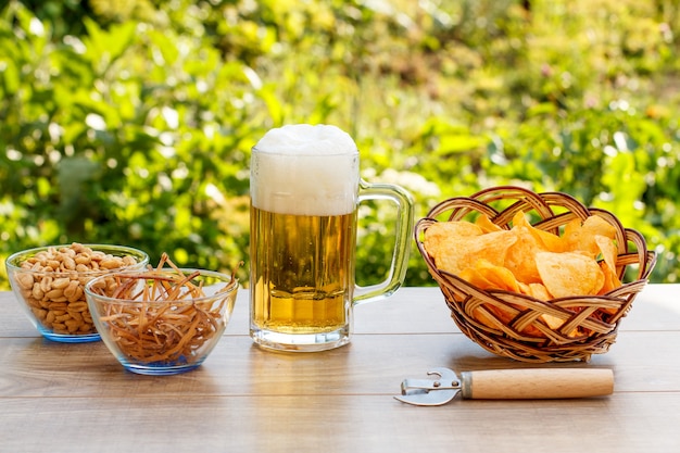 Chope en verre de bière sur une table en bois avec des croustilles dans un panier en osier, des arachides et des calmars séchés dans des bols sur fond flou vert naturel