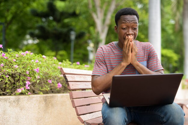 Choqué bel homme africain utilisant un ordinateur portable dans le parc tout en regardant surpris