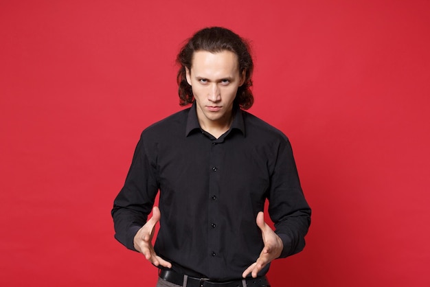 Choqué bouleversé élégant jeune homme aux cheveux longs bouclés en chemise noire posant isolé sur fond de mur rouge portrait en studio. Concept de mode de vie des émotions sincères des gens. Maquette de l'espace de copie. Regarder la caméra.