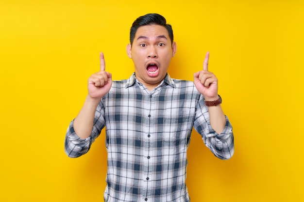 Choqué jeune bel homme asiatique portant une chemise à carreaux pointant le doigt vers l'espace de copie vide présentant un nouveau produit isolé sur fond jaune Concept de style de vie des gens