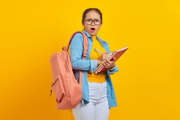 Choqué jeune étudiante asiatique en vêtements denim avec sac à dos écrivant des notes dans un cahier isolé sur fond jaune Éducation au concept de collège universitaire de lycée