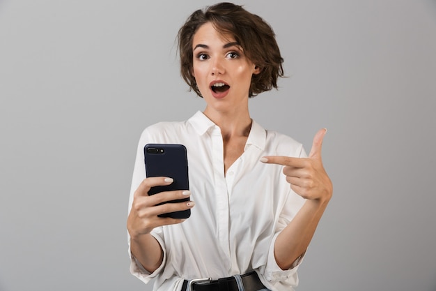 Choqué jeune femme d'affaires posant isolé sur un mur gris à l'aide de téléphone mobile et bavarder