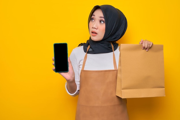 Choqué jeune femme asiatique barista en tablier de t-shirt blanc travail dans un café tenir un sac à emporter en papier vierge maquette et montrant un téléphone portable vierge isolé sur fond jaune
