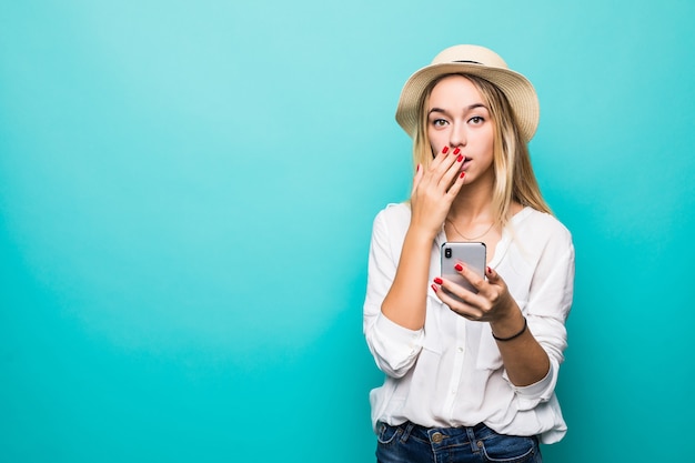 Choqué jeune femme au chapeau de paille à l'aide de téléphone mobile isolé sur mur bleu