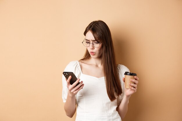 Choqué jeune femme lisant l'écran du téléphone mobile et fronçant les sourcils, tenant une tasse de café, debout sur beige.