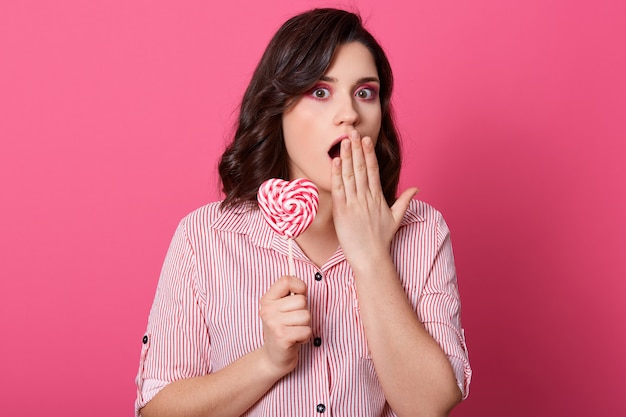 Choqué de jeune femme en vêtements décontractés en gardant la bouche grande ouverte et en la couvrant avec la paume, tenant la sucette