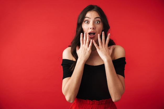 Choqué jeune femme vêtue d'une robe debout isolé sur rouge, bouche ouverte