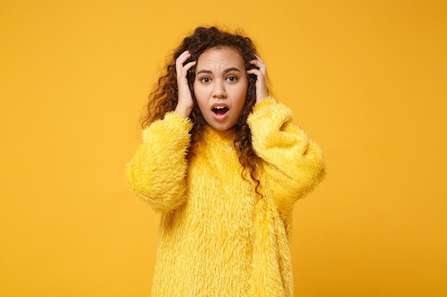 Choqué jeune fille afro-américaine en pull de fourrure posant isolé sur fond de mur orange jaune en studio. Concept de style de vie des gens. Maquette de l'espace de copie. Mettre les mains sur la tête, garder la bouche ouverte.