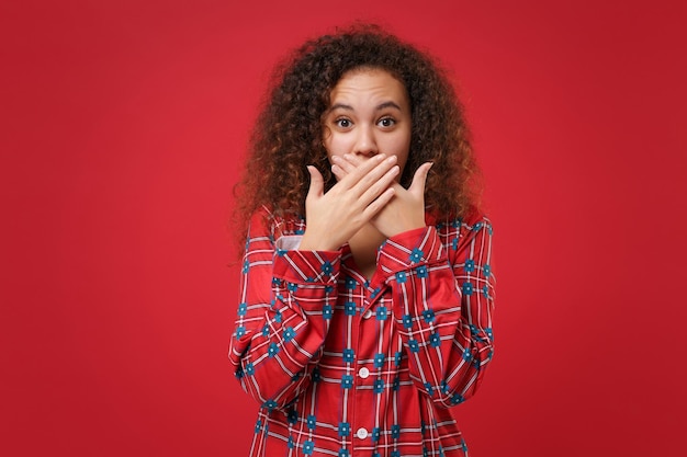 Photo choqué jeune fille afro-américaine en pyjama homewear posant tout en se reposant à la maison isolé sur fond de mur rouge. détendez-vous le concept de mode de vie de bonne humeur. maquette de l'espace de copie. se couvrir la bouche avec les mains.