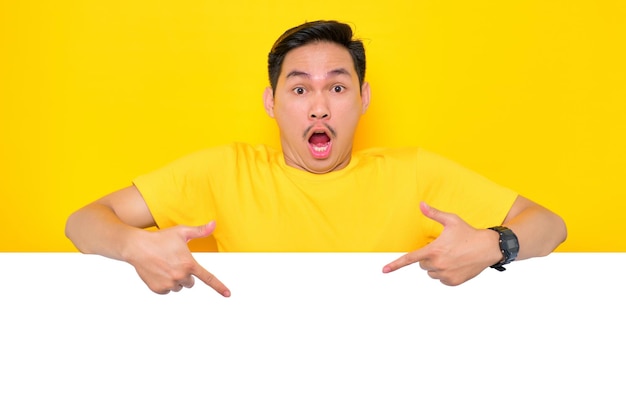 Choqué jeune homme asiatique en t-shirt décontracté pointant vers le bas au panneau publicitaire blanc isolé sur fond jaune Concept de panneau d'affichage de promotion
