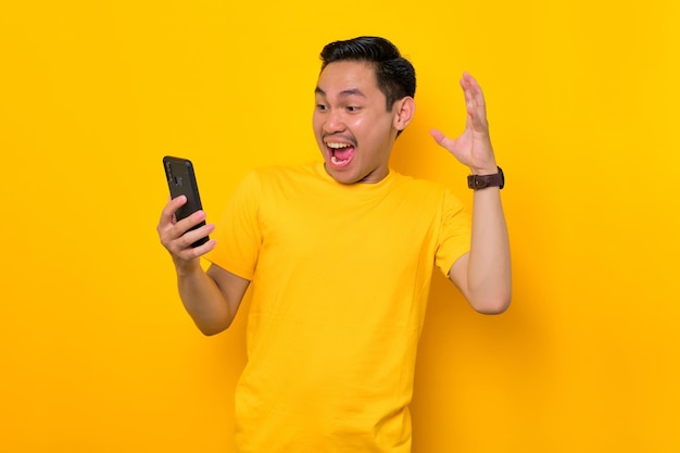 Choqué jeune homme asiatique en t-shirt décontracté regardant l'écran du téléphone portable réagissant aux nouvelles en ligne isolé sur fond jaune Concept de style de vie des gens