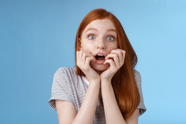 Choqué sans voix haletant jeune fille rousse regardant impressionné stupéfait en regardant la série télévisée moment important mordre les doigts bouche ouverte secoué debout excité fond bleu anticipant.