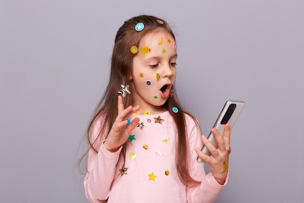 Choqué surpris petite fille couverte d'autocollants colorés pour enfants isolés sur fond gris tenant un téléphone portable dans les mains lisant des nouvelles choquantes sur Internet