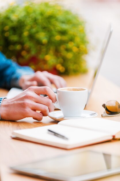 Choses pour le succès. Gros plan sur un jeune homme travaillant sur un ordinateur portable et tenant la main sur une tasse de café