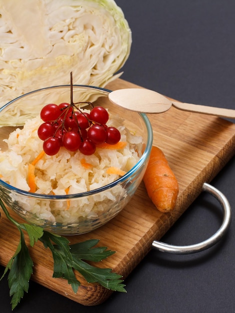 Chou fermenté maison avec carotte, grappe de viorne dans un bol en verre et cuillère en bois. Tête fraîche de chou sur le fond. Salade végétalienne. Le plat est riche en vitamine U. Aliments excellents pour une bonne santé.