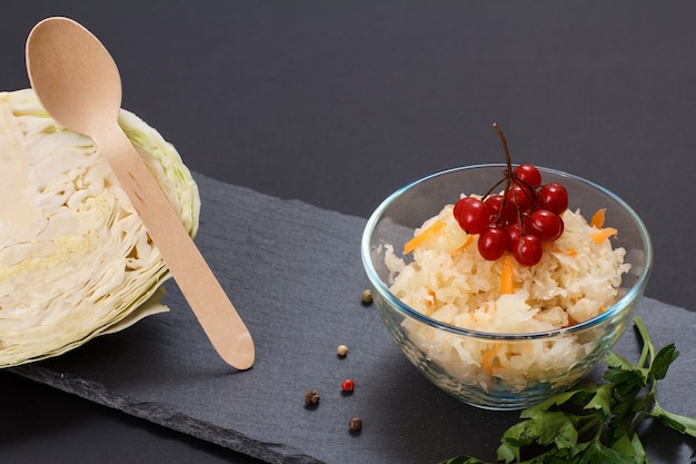 Chou Fermenté Maison Avec Carotte Et Grappe De Viorne Dans Un Bol En Verre Sur Planche De Pierre. Tête Fraîche De Chou Et Cuillère En Bois Sur Le Fond. Salade Végétalienne. Le Plat Est Riche En Vitamine U.