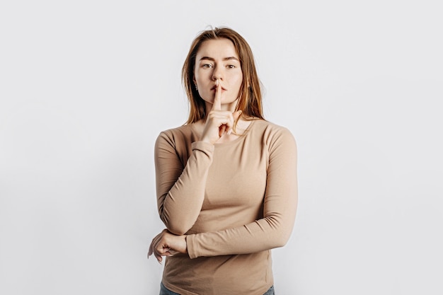 Chut geste. Jeune belle fille sérieuse tenant un doigt sur ses lèvres sur un fond blanc isolé. Une femme demande à se taire, un lieu de publicité. Brune négative dans un pull beige.