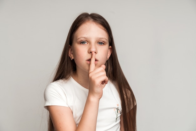 Chut, tais-toi ! Portrait d'une drôle de petite fille mignonne faisant un geste de silence avec le doigt sur ses lèvres, gardant un secret, un mystère d'enfant. Prise de vue en studio isolé sur fond blanc