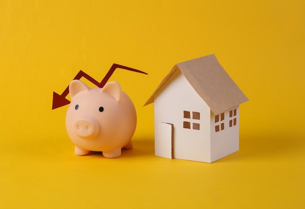 Chute des prix des logements Maison figurine et tirelire avec flèche tombante sur fond jaune