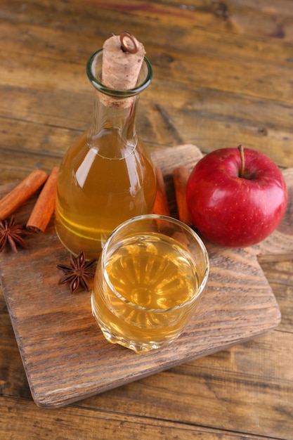 Cidre de pomme dans une bouteille en verre avec des bâtons de cannelle et des pommes fraîches sur une planche à découper, sur un mur en bois