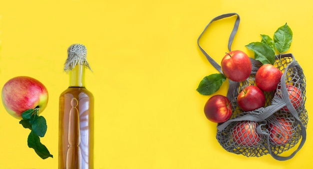 Cidre de vinaigre de pomme dans la bouteille en verre et sac à provisions en filet avec pommes rouges sur fond jaune