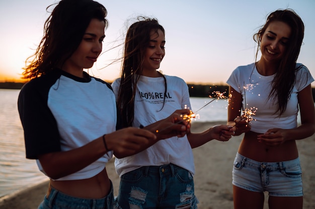 Les cierges magiques entre les mains des jeunes filles sur la plage. Trois filles profitant d'une fête sur la plage avec des cierges magiques. Concept de vacances d'été, vacances, détente et style de vie.