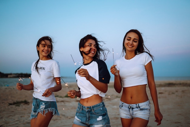 Les cierges magiques entre les mains des jeunes filles sur la plage. Trois filles profitant d'une fête sur la plage avec des cierges magiques. Concept de vacances d'été, vacances, détente et style de vie.