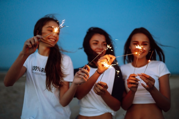 Les cierges magiques entre les mains des jeunes filles sur la plage. Trois filles profitant d'une fête sur la plage avec des cierges magiques. Concept de vacances d'été, vacances, détente et style de vie.