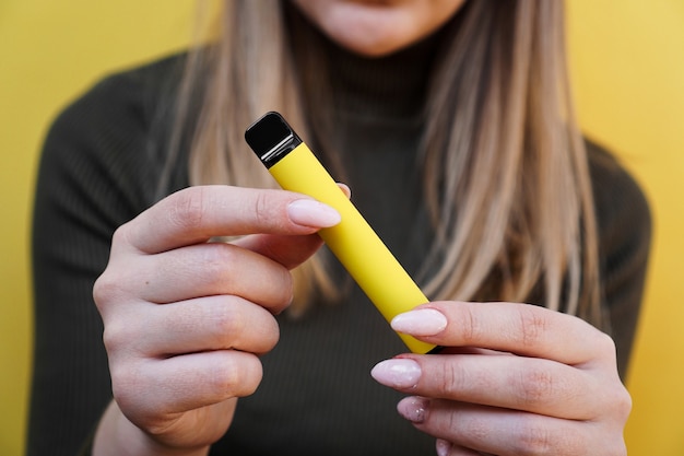 Cigarette électronique jetable jaune dans une main féminine. Fond jaune vif. Vape au melon, ananas ou citron