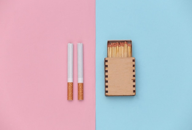 Cigarettes et boîte d'allumettes sur fond pastel rose bleu. Vue de dessus