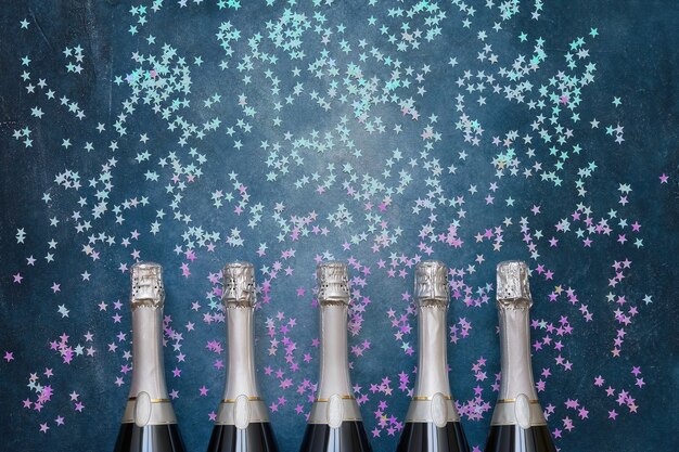 Cinq bouteilles de champagne avec des étoiles de confettis holographiques sur bleu. Copier l'espace, vue de dessus