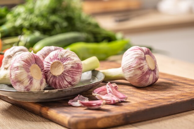 Cinq têtes d'ail sur une planche à découper avec des légumes verts en arrière-plan.