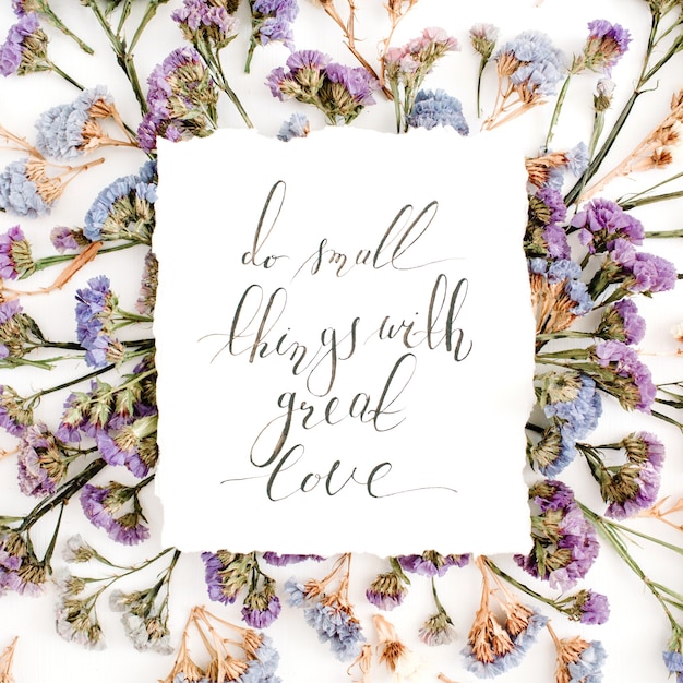 Citation inspirante "Faites de petites choses avec beaucoup d'amour" écrite en style calligraphique sur papier avec des fleurs séchées bleues et violettes sur fond blanc. Mise à plat, vue de dessus