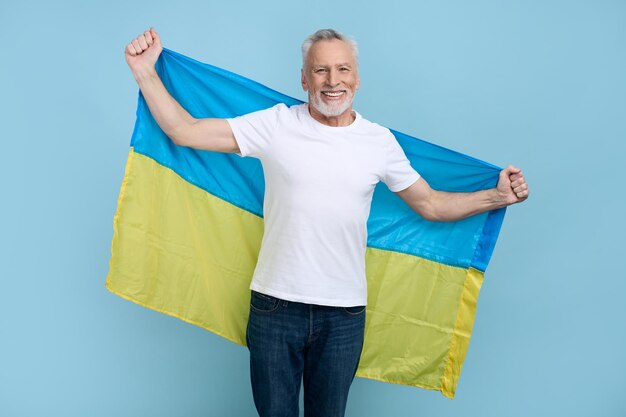 Un citoyen patriotique bénévole senior sourit à la caméra portant le drapeau de l'Ukraine isolé sur bleu