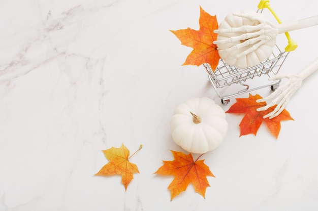 Citrouille d'halloween blanche dans un petit chariot d'épicerie avec des bras squelettes et des feuilles d'érable sur le dos en marbre