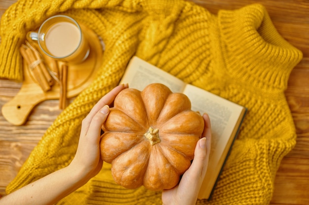 Citrouille mûre dans les mains des femmes sur un latte aromatique de pull tricoté avec des bâtons de cannelle sur un plateau en bois...