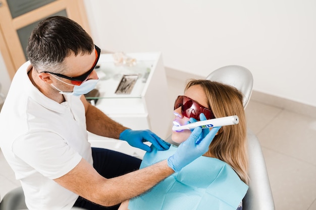 Éclairage uv de l'obturation dentaire photopolymère pour femme en dentisterie Dentiste dans des lunettes de protection rouges traite et élimine les caries d'une patiente