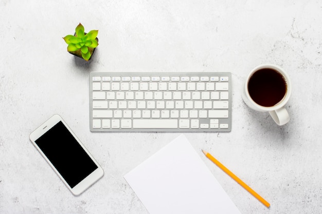 Clavier, une feuille de papier vierge et un crayon, un téléphone, une tasse de café et une plante d'intérieur sur un fond en béton.