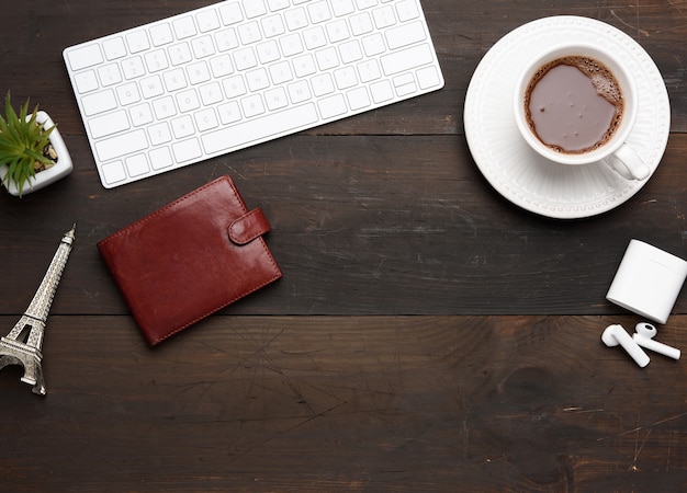 Clavier sans fil blanc et écouteurs sans fil et portefeuille en cuir marron sur table marron en bois, à côté d'une tasse blanche avec café, vue du dessus