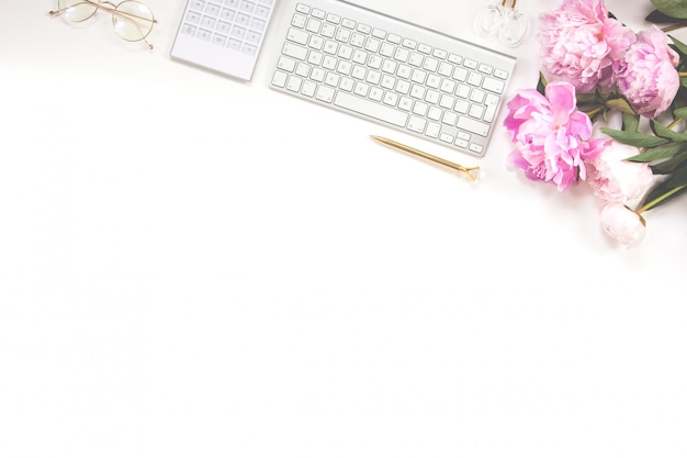 Clavier, stylo doré, lunettes, calculatrice et un bouquet de pivoines roses sur fond blanc. Copiez l'espace.