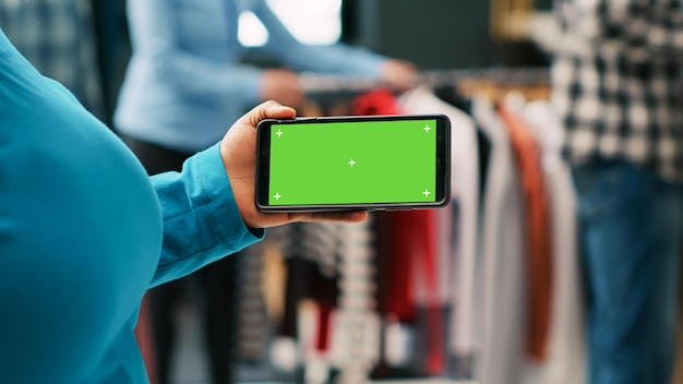 Client afro-américain regardant un téléphone avec un modèle d'écran vert, analysant un affichage isolé dans une boutique moderne. Femme élégante faisant du shopping pour des vêtements décontractés dans un magasin de vêtements. Notion de mode